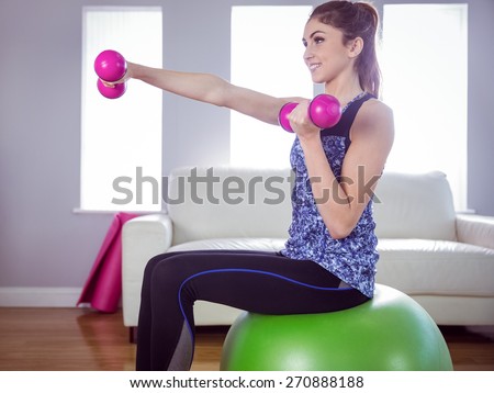 Fit woman lifting dumbbells on exercise ball at home in the living room