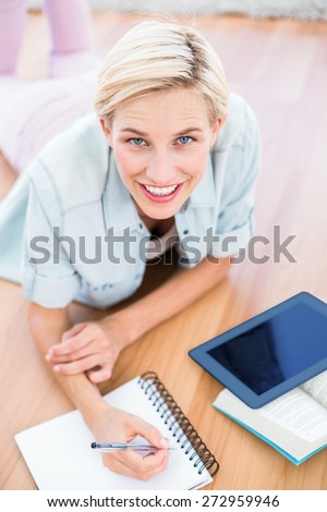 Pretty blonde woman lying on the floor and taking notes while using her tablet in the living room