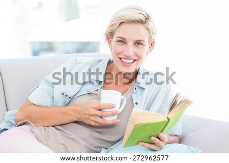 Pretty blonde woman reading a book and holding a mug in the living room