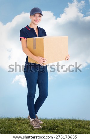 Happy delivery woman holding cardboard box against blue sky over green field