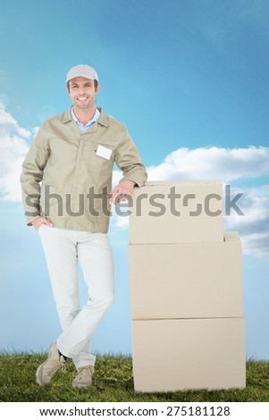 Confident delivery man standing by stack of boxes against blue sky over green field