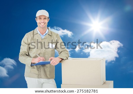 Happy delivery man writing on clipboard by cardboard boxes against bright blue sky with clouds