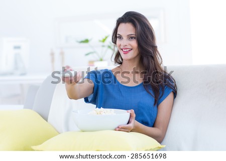 Smiling beautiful brunette relaxing on the couch with bowl of popcorn and changing tv station in the living room