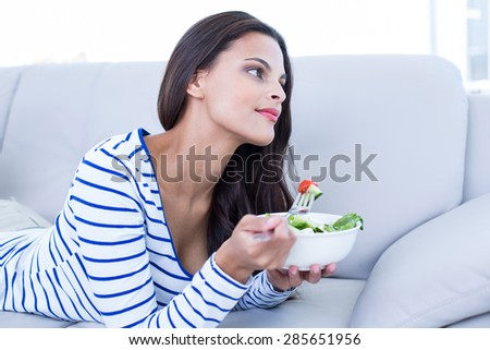 Smiling beautiful brunette relaxing on the couch and eating salad in the living room