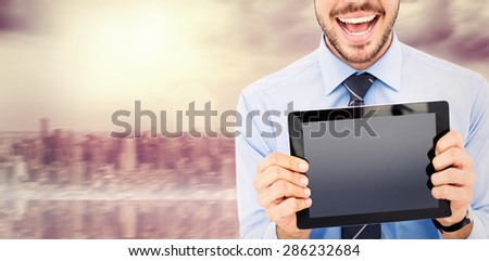 Happy businessman showing his tablet pc against room with large window looking on city