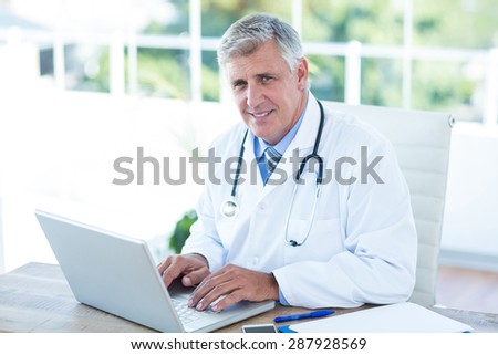 Smiling doctor working on laptop at his desk in medical office