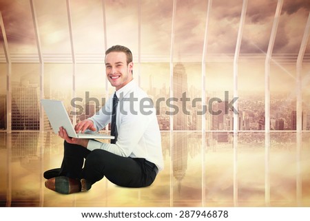 Happy businessman sitting and using laptop against room with large window looking on city