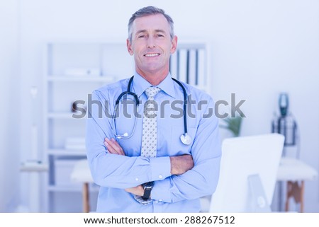 Happy male doctor looking at camera with arms crossed in medical office