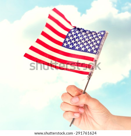 Hand waving american flag against blue sky over green field