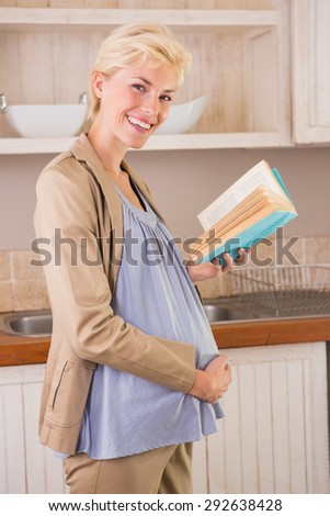 Blonde pregnancy smiling at camera and take a book in the kitchen