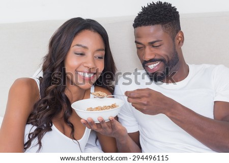 Relaxed couple in bed together eating cereal at home in bedroom