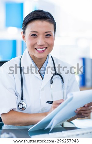 Happy doctor writing on a clipboard and smiling at camera in medical office