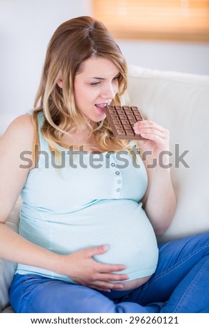 Pretty pregnant woman eating big bar of chocolate at home in the living room