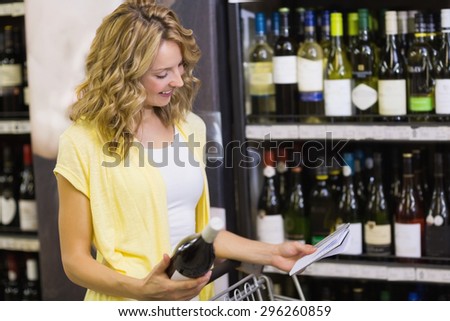 Smiling pretty blonde woman looking at notepad amd having in her hands a wine bottle in supermarket