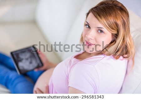 Pregnant woman looking at camera with hand on belly and ultrasound scans in the living room