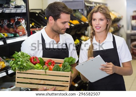 Two colleagues holding a box with fresh vegetables and writing on notepad at supermarket