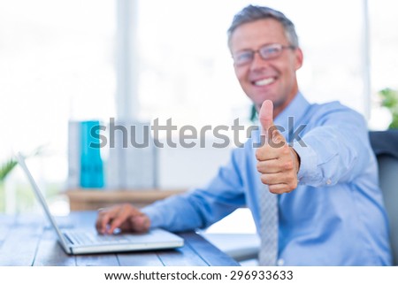 Happy businessman using laptop computer and looking at camera with thumbs up in office