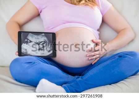 Pregnant woman showing ultrasound scans and touching her belly in the living room