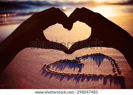 Hands making heart shape on the beach against one heart drawn in the sand