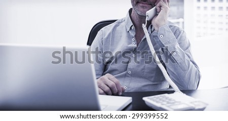 Serious mature businessman on call in front of laptop at desk in a bright office