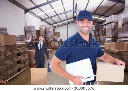 Happy delivery man with cardboard box and clipboard against forklift in a large warehouse