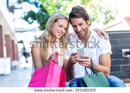 Smiling couple with shopping bags sitting and using smartphone at shopping mall