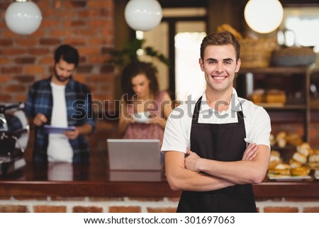 Portrait of smiling barista with arms crossed in front of customers at coffee shop