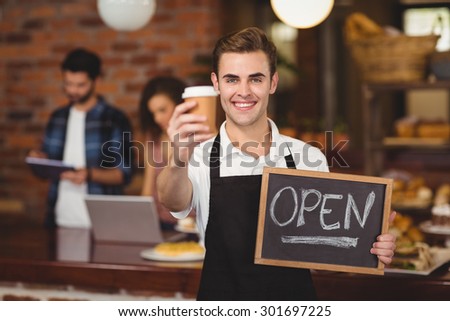 Portrait of smiling barista holding take-away cup and open sign at coffee shop