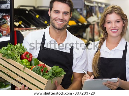 Portrait of two colleagues holding a box with fresh vegetables and writing on notepad at supermarket
