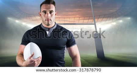 Tough rugby player holding ball against rugby stadium