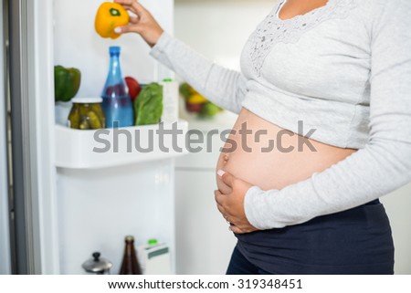 Midsection of pregnant woman holding belly pepper from fridge while touching her belly in kitchen