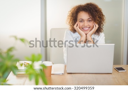 Portrait of smiling businesswoman with hand on chin sitting by laptop on desk in office