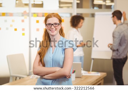 Portrait of confident businesswoman with arm crossed sitting on table with colleagues looking at whiteboard in background at office