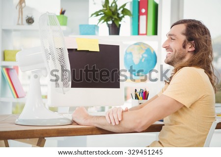 Happy hipster sitting by electric fan on computer desk in office
