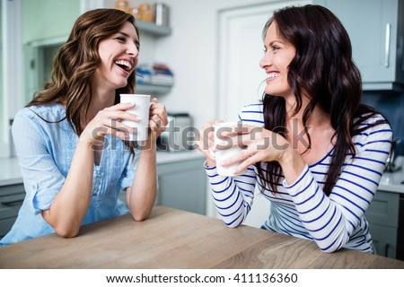 Happy female friends holding coffee mugs while discussing at table in kitchen