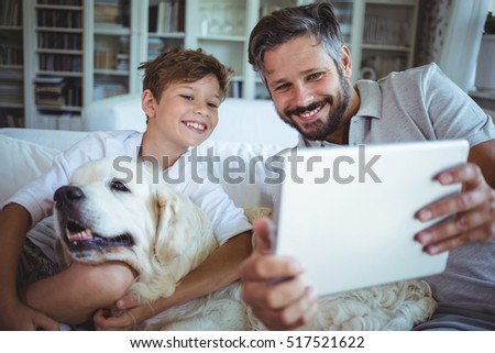 Father and son sitting on sofa with pet dog and using digital tablet in living room at home