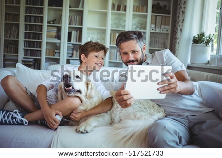 Father and son sitting on sofa with pet dog and using digital tablet in living room at home
