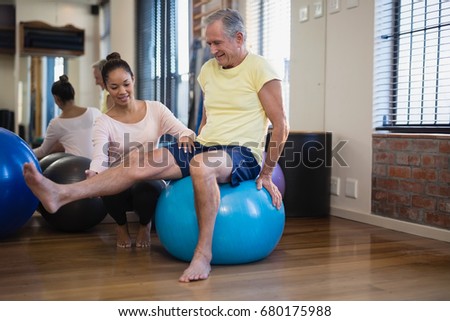 Female therapist helping senior male patient doing leg exercise on blue ball at hospital ward