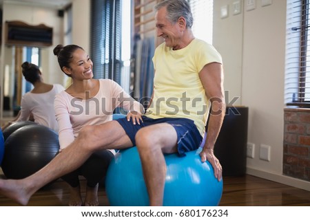 Smiling female therapist crouching by senior male patient sitting on exercise ball at hospital ward