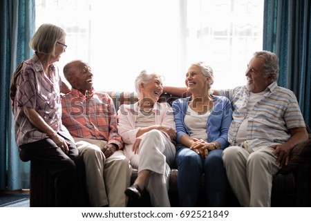 Smiling senior friends talking while sitting on couch against window in nursing home