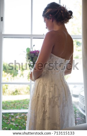 Side view of bride holding bouquet while looking through window at home
