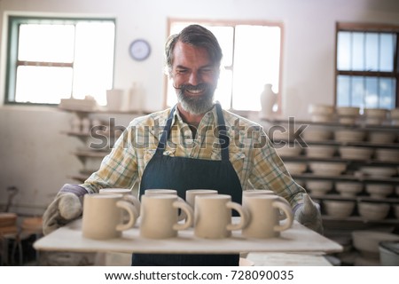 Happy male potter holding ceramic cups in tray at pottery workshop