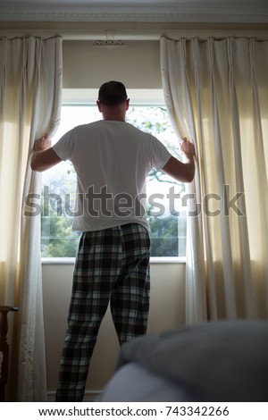 Rear view of man opening window curtain in bedroom