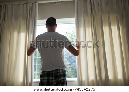 Rear view of man opening window curtain in bedroom