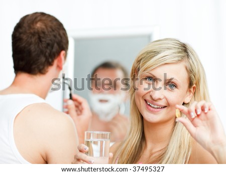 Smiling woman holding pills and man shaving in bathroom