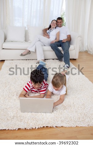 Brother and sister using a laptop on floor in living-room with their parents on sofa