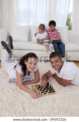 Couple playing chess on floor in living-room with their children on sofa