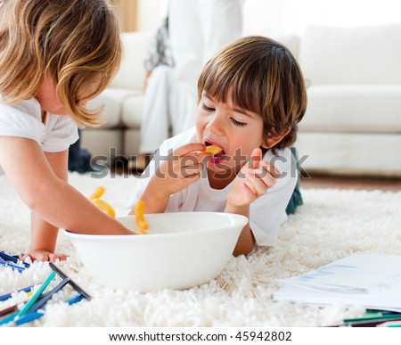 Joyful siblings eating chips and drawing lying on the floor