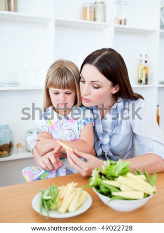 Dissatisfied little girl eating vegetables with her mother in the kitchen