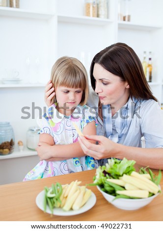 Annoyed blond girl eating vegetables with her mother in the kitchen